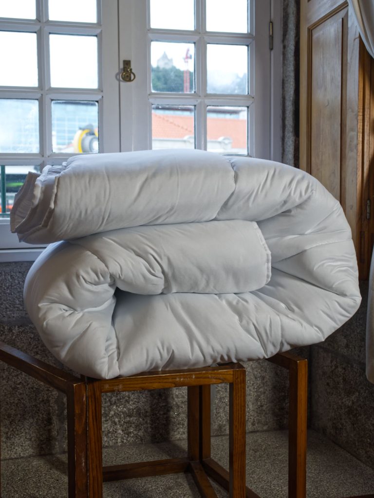 Edredão nórdico branco de 400g dobrado e apoiado numa cadeira de madeira junto a uma janela.