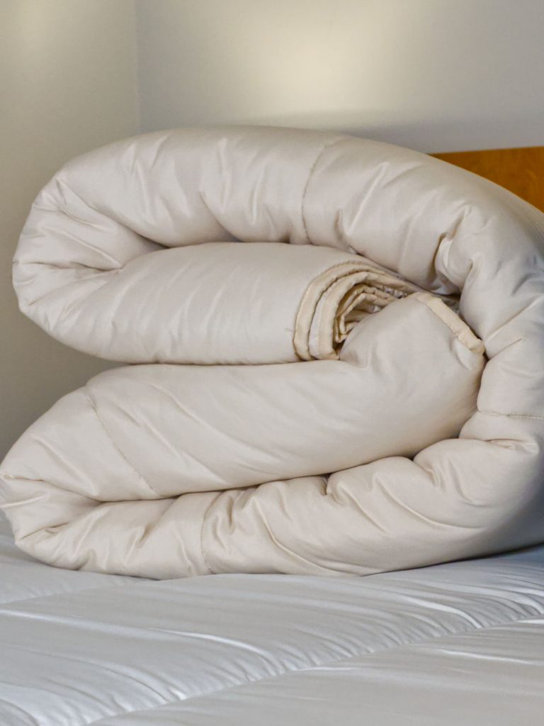 Edredão nórdico bege de 200g enrolado sobre a cama, exibindo a espessura do enchimento.