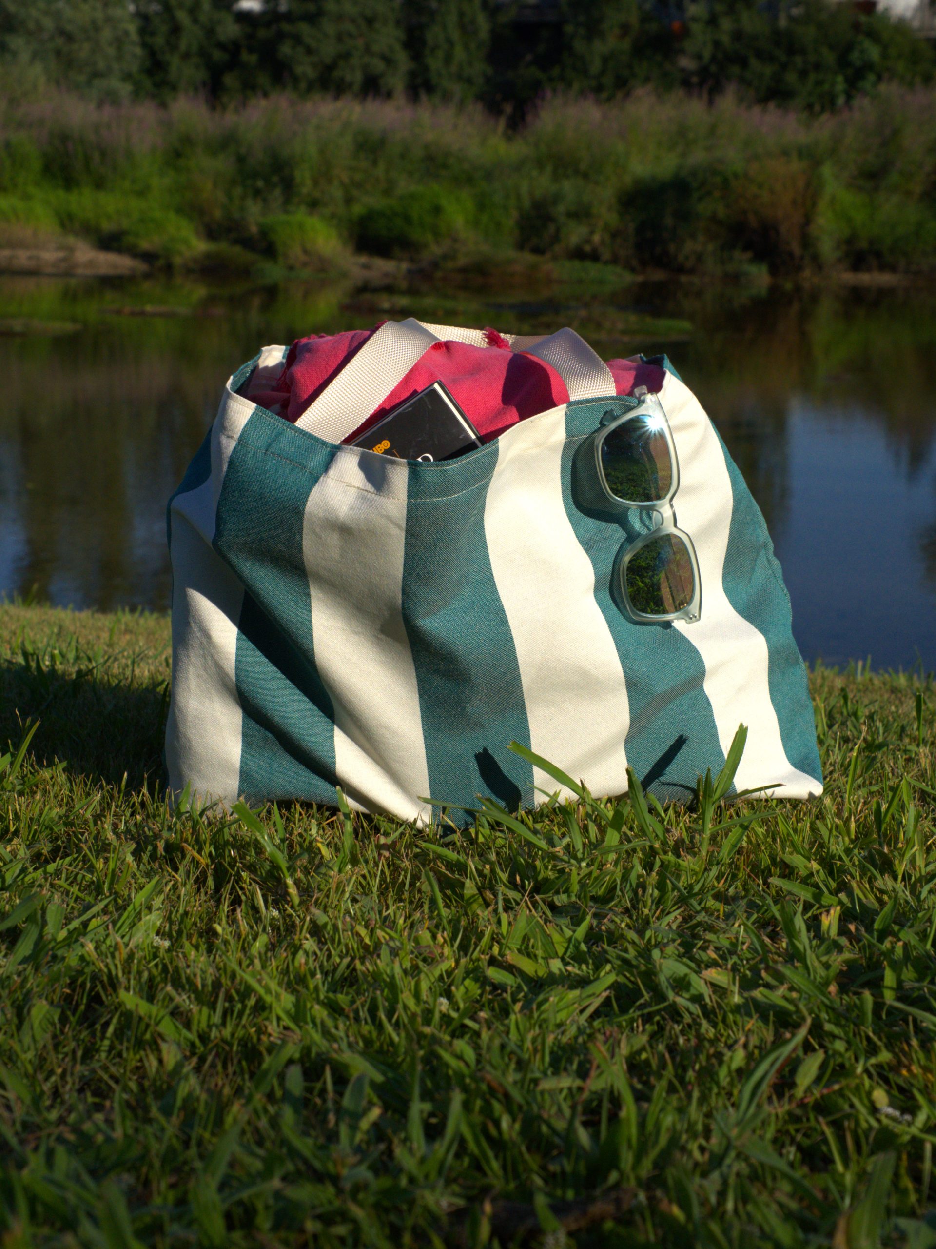 Saco de praia LeCamaleon com padrão às riscas verdes e brancas, tecido em poliéster leve, mostrado em uso ao ar livre perto de um lago. Ideal para atividades de verão e uso sustentável.